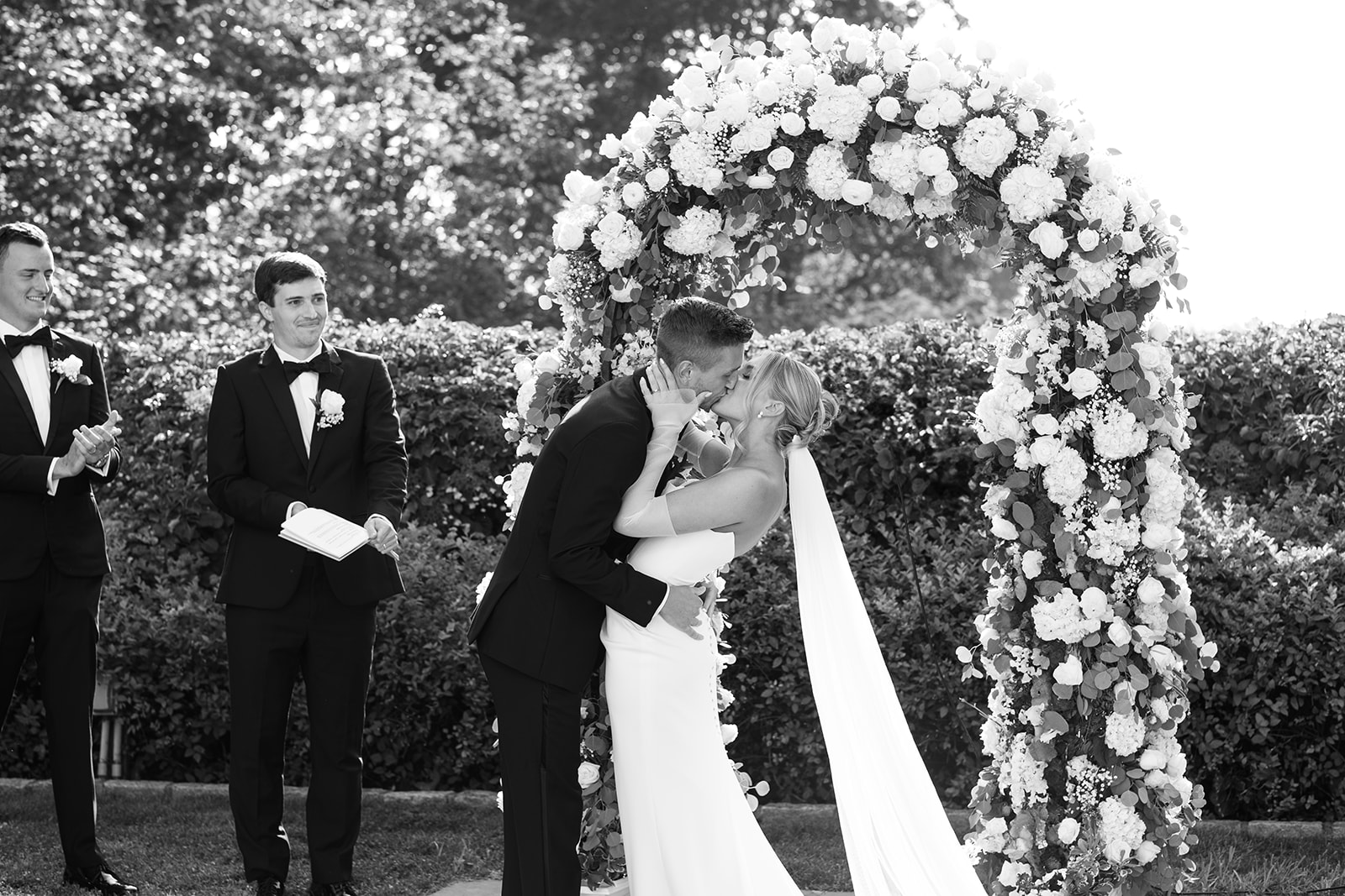 Black and white photo of the groom dipping and kissing the bride under a lush floral arch.